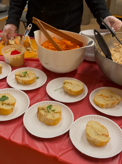 To show part of the set up of the table at which the Vietnamese Banh Mi Bites were being assembled and served at. There are several plates of slices of the sandwhich, infront two bowls. One bowl is filled with pickled carrots and the other with crumbled tofu.